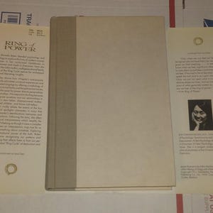 K&ouml;nnte beinhalten: Ein gebundenes Buch mit dem Titel Ring of Power von Jean Shinoda Bolen, das einen schlichten hellbeigen Einband und einen grauen Buchr&uuml;cken hat. Das Buch liegt mit ausgebreiteten Schutzumschlagklappen da, die Text, ein Autorenfoto und Logos zeigen.
