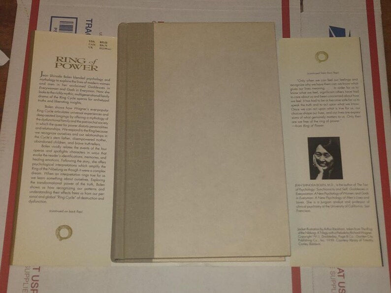 K&ouml;nnte beinhalten: Ein gebundenes Buch mit dem Titel Ring of Power von Jean Shinoda Bolen, das einen schlichten hellbeigen Einband und einen grauen Buchr&uuml;cken hat. Das Buch liegt mit ausgebreiteten Schutzumschlagklappen da, die Text, ein Autorenfoto und Logos zeigen.