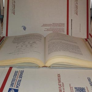 Puede incluir: Un libro abierto con p&aacute;ginas amarillentas, probablemente antiguo, apoyado sobre una superficie de cajas USPS. El texto es visible, lo que sugiere que es un manual o libro de referencia. El lomo del libro es visible y las p&aacute;ginas est&aacute;n ligeramente curvadas.