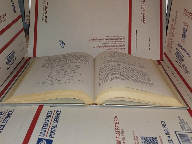 Puede incluir: Un libro abierto con p&aacute;ginas amarillentas, probablemente antiguo, apoyado sobre una superficie de cajas USPS. El texto es visible, lo que sugiere que es un manual o libro de referencia. El lomo del libro es visible y las p&aacute;ginas est&aacute;n ligeramente curvadas.