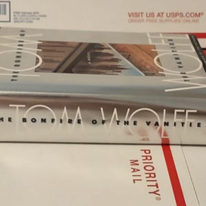 May include: A hardcover book titled "The Bonfire of the Vanities" by Tom Wolfe. The book has a silver cover with the title in large white letters. The book is lying on a white and red Priority Mail package.