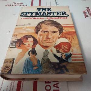 May include: The Spymaster, a novel of America by Donald Freed, is the title of this paperback book with a colorful cover featuring a man, a woman, and an American flag.