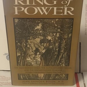 K&ouml;nnte beinhalten: Ein gebundenes Buch mit dem Titel Ring of Power von Jean Shinoda Bolen, M.D. Der goldene Einband zeigt eine schwarz-wei&szlig;e Illustration einer Person mit einem Pferd in einem Wald. Der Untertitel lautet The Abandoned Child, The Authoritarian Father, And The Disempowered Feminine, A Jungian Understanding of Wagner's Ring Cycle.