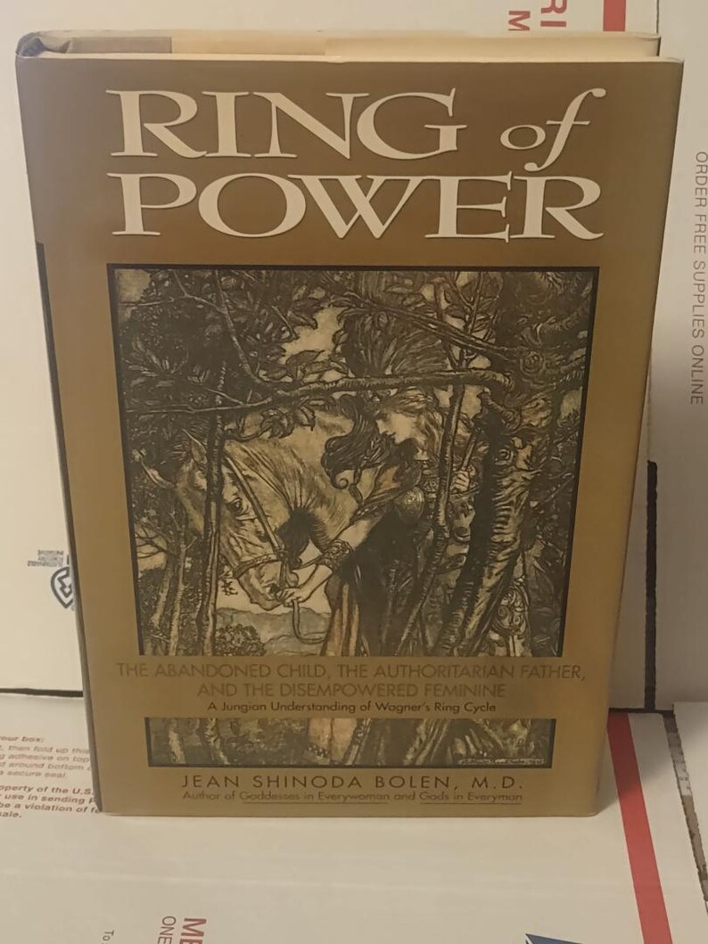 K&ouml;nnte beinhalten: Ein gebundenes Buch mit dem Titel Ring of Power von Jean Shinoda Bolen, M.D. Der goldene Einband zeigt eine schwarz-wei&szlig;e Illustration einer Person mit einem Pferd in einem Wald. Der Untertitel lautet The Abandoned Child, The Authoritarian Father, And The Disempowered Feminine, A Jungian Understanding of Wagner's Ring Cycle.