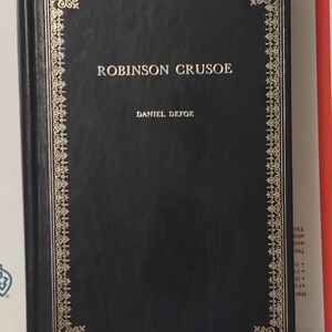 May include: A hardcover book titled "ROBINSON CRUSOE" by Daniel Defoe. The book has a dark blue cover with gold decorative border and lettering. The spine is also dark blue.