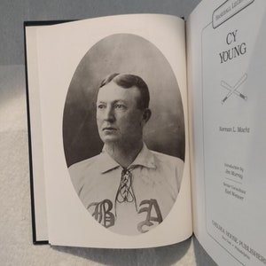 May include: A black and white photo of Cy Young, a baseball legend, wearing a baseball uniform with the letters "BA" on the front. The photo is from the book "Baseball Legends: Cy Young" by Norman L. Macht.