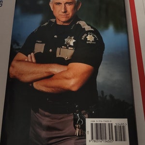 May include: A close-up of a man in a black and tan sheriff's uniform with the words "King County Sheriff" on the patch. He is standing with his arms crossed and looking directly at the camera. The background is blurred and appears to be a lake or river.