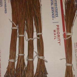 May include: Bundles of dried pine needles tied with white twine. The needles are brown with some green tips. The bundles are arranged on a white and blue USPS Priority Mail box.