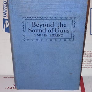 May include: A vintage book titled "Beyond the Sound of Guns" by Emilie Loring. The book has a light blue cover with the title and author's name in dark blue lettering within a decorative frame. The book is slightly worn.