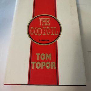 May include: A white book with a red spine and a black circle with gold lettering that reads "THE CODICIL A NOVEL" and "TOM TOPOR" in gold lettering.