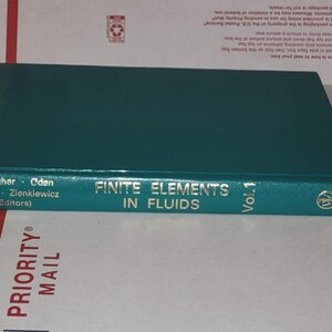 May include: A teal hardcover book titled "Finite Elements in Fluids Vol. 1" by Wiley. The book's spine features the names of the editors and the Wiley logo. The book is lying on a white and brown surface.