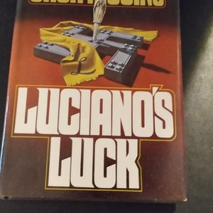 May include: A brown book cover with the title "Luciano's Luck" in white letters. The author's name, "Jack Higgins", is printed in yellow letters above the title. The book cover features a yellow cloth with a cross-shaped design and a small, silver knife.