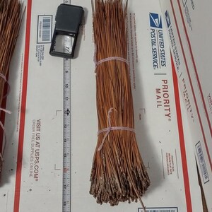 May include: A bundle of long, dried brown pine needles tied with a light-colored string. A metal measuring tape is positioned next to the bundle, showing a length of approximately 15 inches from the base to the tips of the needles.