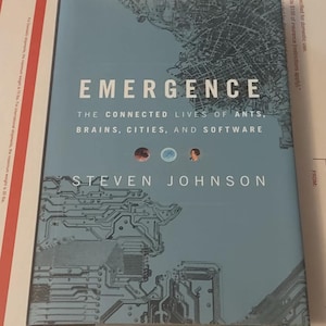 May include: A hardcover book titled "Emergence" by Steven Johnson. The cover is light blue with a city map design and circuit board graphics. The book's title and author's name are prominently displayed.