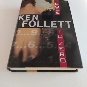 May include: A paperback book titled "Code to Zero" by Ken Follett. The cover features a blurry image of a person walking down a street with numbers in the background.