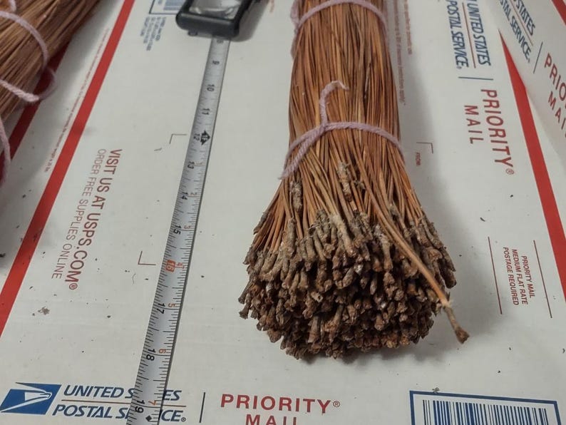 May include: A bundle of dried pine needles, tied with pink twine. A metal ruler is placed next to the bundle, showing measurements in inches. The bundle is on a white surface with USPS Priority Mail branding.