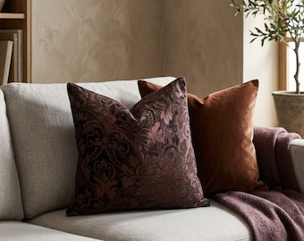 May include: Two square throw cushions placed on a light grey sofa. The front cushion is a dark brown velvet with a raised, tonal damask pattern. The back cushion is a solid, smooth, medium-brown velvet. A dark plum-coloured throw blanket is draped nearby.