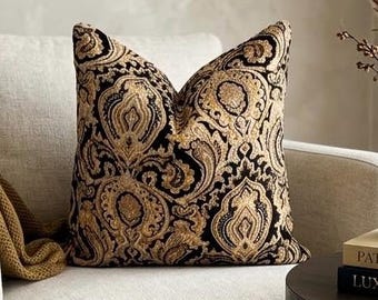May include: A square throw pillow featuring a prominent black and gold paisley pattern. The fabric displays intricate, swirling ornamental motifs in metallic gold thread set against a deep black background, creating a high-contrast, textured appearance.