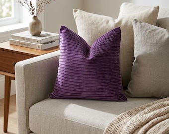 May include: A square throw cushion featuring a deep purple colour and a horizontal ribbed texture. The cushion is positioned on a light beige sofa alongside two plain beige throw cushions and a textured beige knit throw blanket draped over the seat.