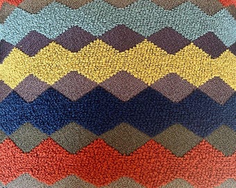 May include: Close-up of a colorful, textured fabric with a repeating zigzag pattern. The design features rows of various colors, including red, blue, yellow, and purple. The fabric has a plush, tactile appearance, suggesting a soft material.
