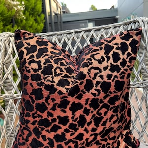 May include: A square cushion with a leopard print design. The cushion has a black pattern on a peach-coloured background. The cushion is placed on a white wicker chair, with greenery visible in the background.