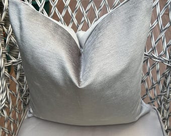 May include: A light gray decorative pillow with a textured surface rests on a matching gray cushion. The pillow is placed on a gray wicker chair, creating a cohesive and elegant look. The pillow's fabric has a subtle sheen.