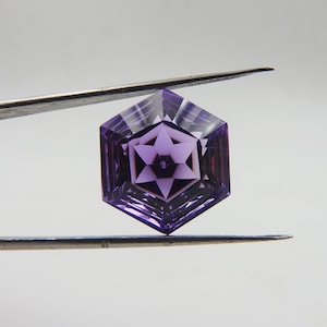 Könnte beinhalten: Ein facettierter, sechseckiger Amethyst-Edelstein, der von einer Pinzette gehalten wird. Der Edelstein weist ein sternförmiges Design in der Mitte auf und zeigt verschiedene Lilatöne. Die Facetten erzeugen ein Spiel aus Licht und Schatten.