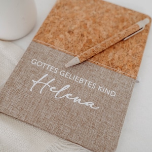May include: A brown cork and fabric journal with the text "Gottes geliebtes Kind" and the name "Helena" written in white. A pen is laying on top of the journal.