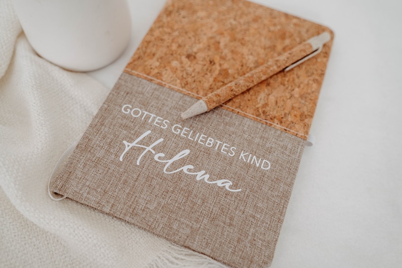 May include: A brown cork and fabric journal with the text "Gottes geliebtes Kind" and the name "Helena" written in white. A pen is laying on top of the journal.