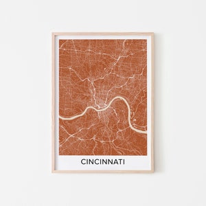 May include: A framed print of a map of Cincinnati, Ohio. The map is in a burnt orange color with white lines showing the streets and roads. The city name is printed in black at the bottom of the map.