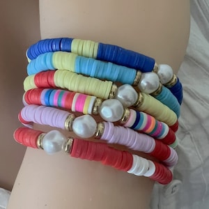 Könnte beinhalten: Ein Stapel bunter Perlenarmbänder mit weißen Perlenakzenten. Die Armbänder bestehen aus runden, flachen Perlen in den Farben Rot, Gelb, Blau, Rosa und Weiß.