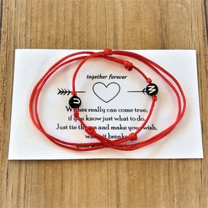 Puede incluir: Dos pulseras de cuerda roja con cuentas negras que tienen las letras "J" y "Z". Las pulseras están atadas juntas con una tarjeta blanca que dice "juntos para siempre" y tiene un corazón dibujado. La tarjeta también tiene una cita sobre los deseos que se hacen realidad.