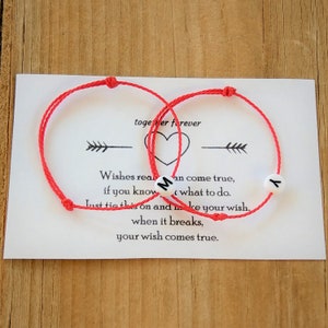 Puede incluir: Dos pulseras de cuerda roja con cuentas blancas que deletrean las letras "M" y "Y". Las pulseras están sobre una tarjeta blanca con el texto "juntos para siempre" y un símbolo de corazón. La tarjeta también incluye instrucciones sobre cómo atar las pulseras y hacer un deseo.