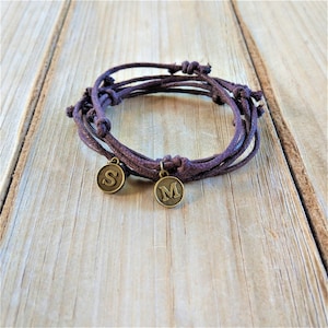 Puede incluir: Un brazalete de cordón marrón con dos abalorios de metal, uno con la letra "S" y el otro con la letra "M".