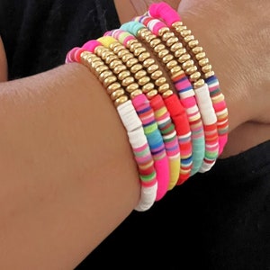 May include: A stack of colorful bracelets with gold beads. The bracelets are made of different colored beads and are stacked on top of each other. The bracelets are on a person's wrist.