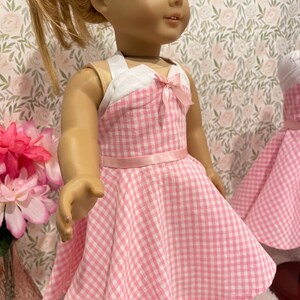 Pode incluir: Uma boneca vestindo um vestido rosa e branco xadrez com corpete branco e la&ccedil;o rosa. O vestido tem uma saia rodada e um cinto de fita rosa. A boneca est&aacute; usando t&ecirc;nis brancos. O fundo apresenta um papel de parede floral.