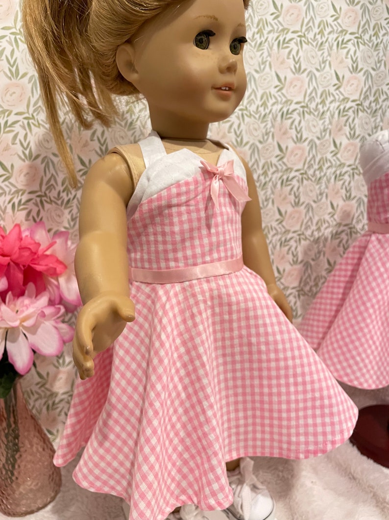 Pode incluir: Uma boneca vestindo um vestido rosa e branco xadrez com corpete branco e la&ccedil;o rosa. O vestido tem uma saia rodada e um cinto de fita rosa. A boneca est&aacute; usando t&ecirc;nis brancos. O fundo apresenta um papel de parede floral.