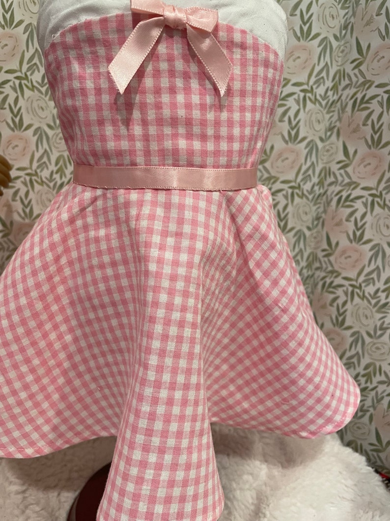 Pode incluir: Um vestido rosa e branco xadrez com um la&ccedil;o de fita rosa e um cinto de fita a condizer. O vestido &eacute; desenhado com um corpete sem al&ccedil;as e uma saia rodada. O tecido tem um padr&atilde;o xadrez cl&aacute;ssico.