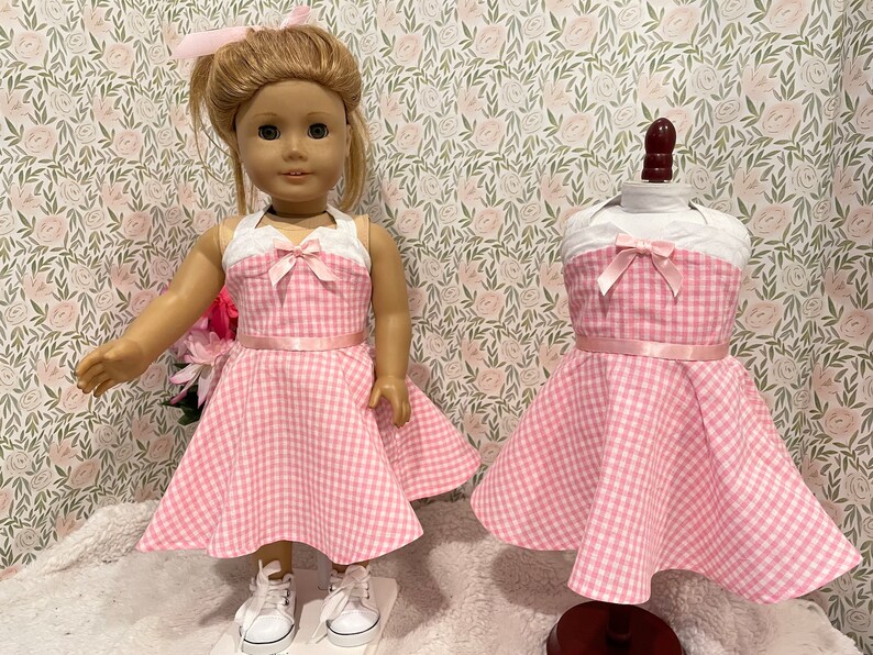 Pode incluir: Um vestido rosa e branco xadrez para boneca, com um top branco de cabresto e um la&ccedil;o rosa. O vestido &eacute; exibido em uma boneca e em um manequim. A boneca est&aacute; usando t&ecirc;nis brancos e tem um la&ccedil;o rosa combinando no cabelo.