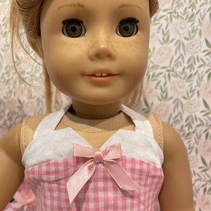 Pode incluir: Uma boneca com cabelo loiro e olhos verdes vestindo um vestido xadrez rosa e branco com um la&ccedil;o rosa. O vestido tem um corpete branco com um padr&atilde;o acolchoado. A boneca tem sardas e est&aacute; sorrindo.