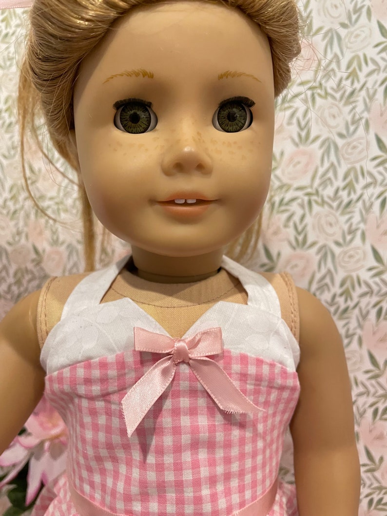 Pode incluir: Uma boneca com cabelo loiro e olhos verdes vestindo um vestido xadrez rosa e branco com um la&ccedil;o rosa. O vestido tem um corpete branco com um padr&atilde;o acolchoado. A boneca tem sardas e est&aacute; sorrindo.
