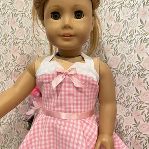 Pode incluir: Boneca vestindo um vestido xadrez rosa e branco com um top halter branco e um la&ccedil;o rosa. A boneca tem cabelo loiro e olhos verdes. Um la&ccedil;o rosa est&aacute; no cabelo da boneca. O fundo &eacute; um padr&atilde;o floral.
