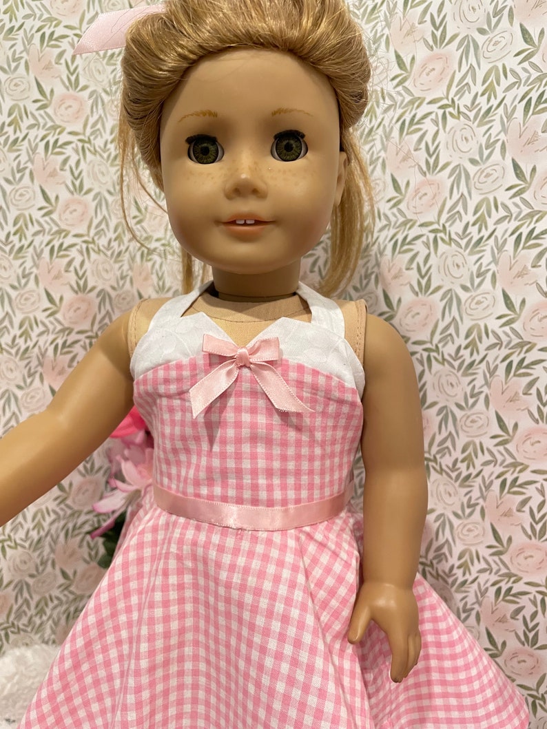 Pode incluir: Boneca vestindo um vestido xadrez rosa e branco com um top halter branco e um la&ccedil;o rosa. A boneca tem cabelo loiro e olhos verdes. Um la&ccedil;o rosa est&aacute; no cabelo da boneca. O fundo &eacute; um padr&atilde;o floral.