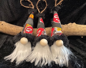May include: Three gnomes with white beards and black hats. The hats are made of fabric with a red, black, and white pattern. The gnomes are hanging from a branch.