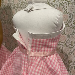 Pode incluir: Um vestido rosa e branco xadrez em um manequim branco. O vestido tem uma gola branca e uma fita rosa na cintura. O vestido &eacute; abotoado nas costas.
