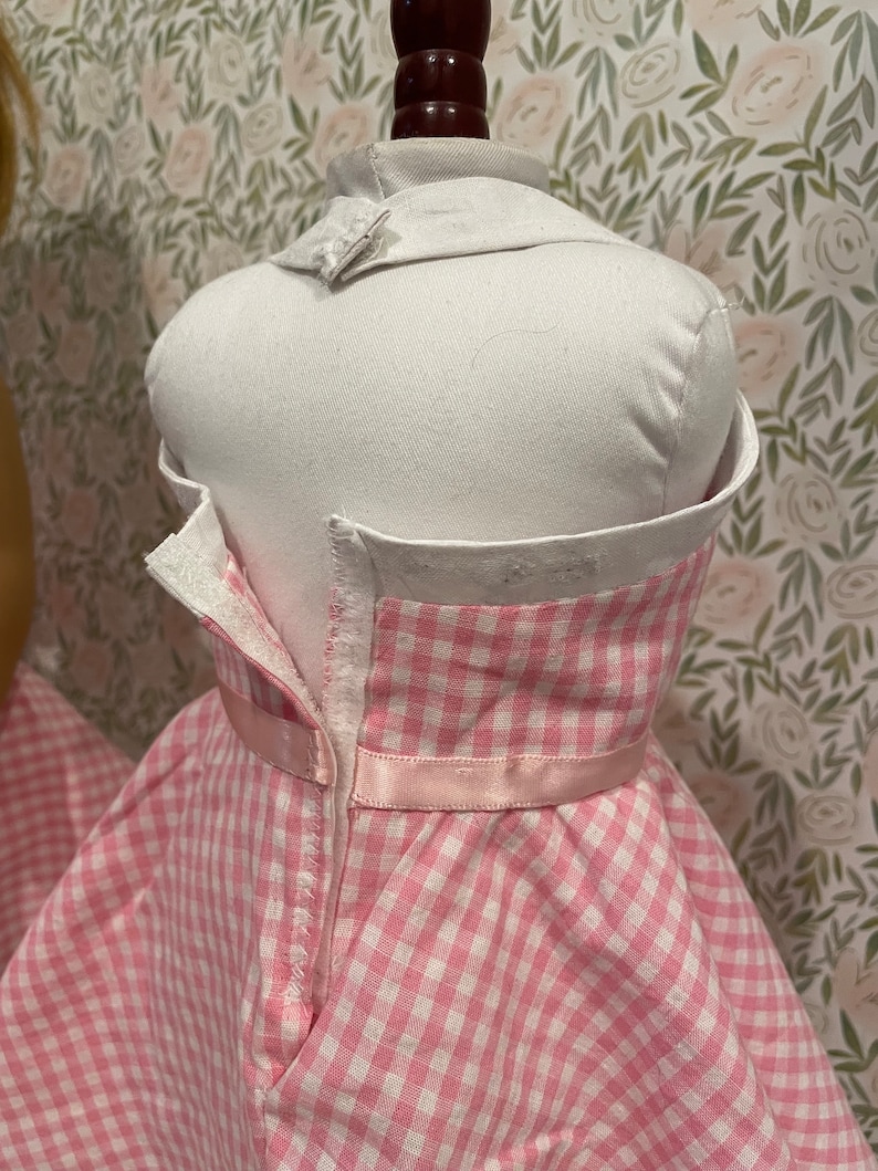 Pode incluir: Um vestido rosa e branco xadrez em um manequim branco. O vestido tem uma gola branca e uma fita rosa na cintura. O vestido &eacute; abotoado nas costas.