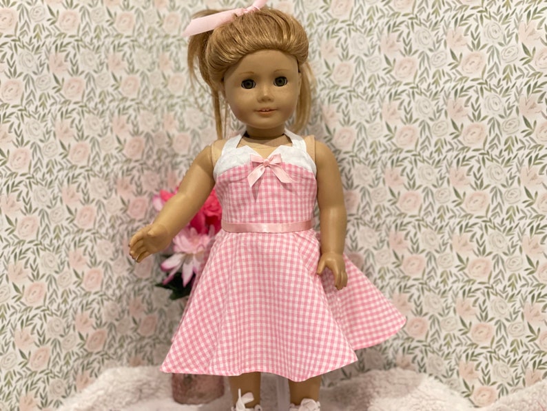 Pode incluir: Uma boneca vestindo um vestido xadrez rosa e branco com um top branco e um la&ccedil;o rosa. O vestido tem um cinto de fita rosa. A boneca tem cabelo loiro preso em um rabo de cavalo com um la&ccedil;o rosa. O fundo &eacute; um papel de parede floral.