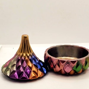 May include: A colorful, geometric trinket box with a lid shaped like a cone. The box is made of ceramic and has a metallic finish. The lid is decorated with a pattern of diamonds in shades of gold, purple, blue, and green.