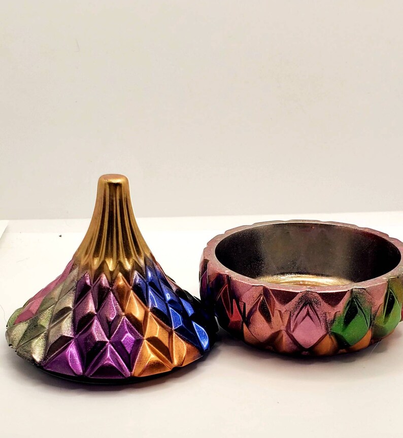 May include: A colorful, geometric trinket box with a lid shaped like a cone. The box is made of ceramic and has a metallic finish. The lid is decorated with a pattern of diamonds in shades of gold, purple, blue, and green.