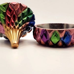 May include: A colorful, geometric trinket box with a lid shaped like a flower. The box is made of resin and has a shiny, metallic finish. The lid is decorated with a pattern of diamonds in shades of pink, red, green, blue, and gold.